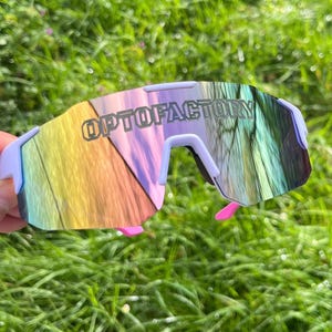 May include: Sport sunglasses with a light purple frame and a mirrored lens that reflects a rainbow of colors. The text "OPTOFACTORY" is printed on the lens. The sunglasses have pink accents on the temples.