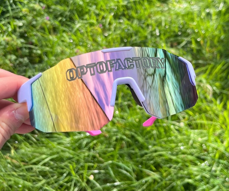 May include: Sport sunglasses with a light purple frame and a mirrored lens that reflects a rainbow of colors. The text "OPTOFACTORY" is printed on the lens. The sunglasses have pink accents on the temples.