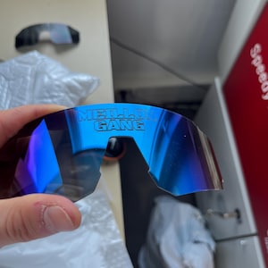 May include: Blue mirrored sunglasses with the text "MEILLO GANG" printed on the lens. The sunglasses have a curved design and a dark frame. The lenses reflect a gradient of blue and purple.
