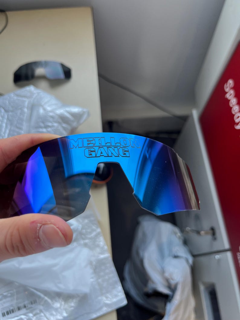 May include: Blue mirrored sunglasses with the text "MEILLO GANG" printed on the lens. The sunglasses have a curved design and a dark frame. The lenses reflect a gradient of blue and purple.