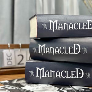 May include: A stack of three hardcover books with the title "Manacled" in white lettering. The books are dark blue with white text and a decorative star design. The book edges are white and red.