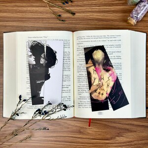 May include: Open book with two black and white character art bookmarks. One bookmark features a side profile of two characters, the other shows a figure with pink accents. A sprig of greenery rests on the book.