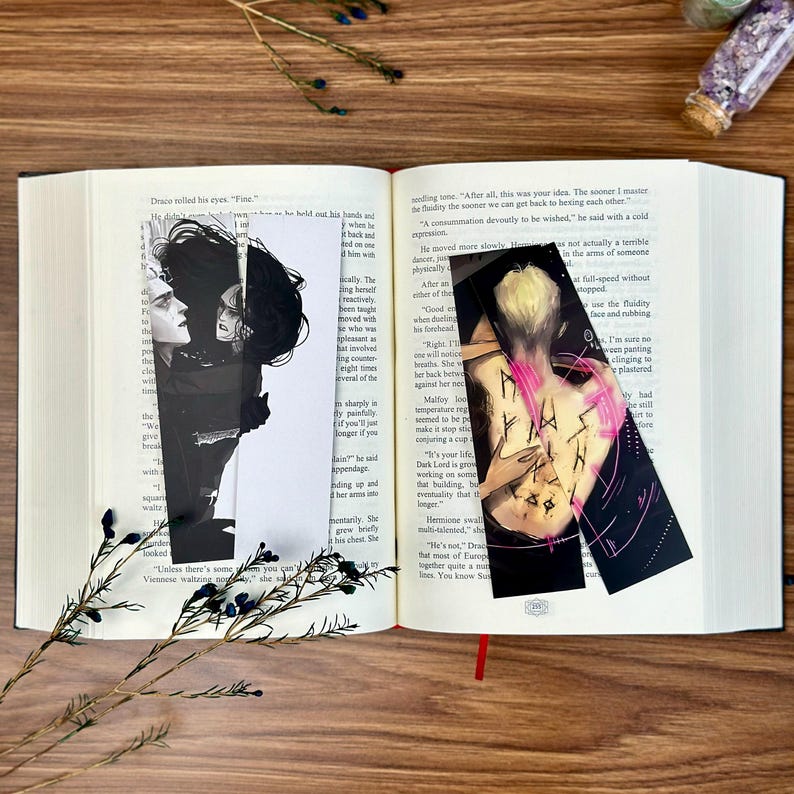 May include: Open book with two black and white character art bookmarks. One bookmark features a side profile of two characters, the other shows a figure with pink accents. A sprig of greenery rests on the book.