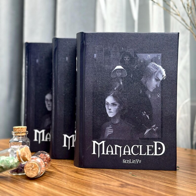 May include: Three dark-colored books with the title "Manacled" in white lettering. The book covers feature illustrations of people in a grayscale palette. Small glass bottles with cork stoppers are in the foreground.