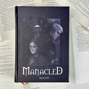 May include: A hardcover book titled "Manacled" by SenLinYu. The cover features a grayscale illustration of several figures with a gothic aesthetic. The book is black with white lettering.