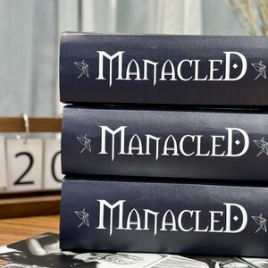 May include: Three dark blue books stacked on top of each other. The word "Manacled" is printed in white on the spine of each book, with a star-like design on either side. The books are on a wooden surface.