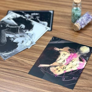 May include: A collection of art prints featuring black and white illustrations and a color print with a figure and symbols. Two small glass bottles with cork stoppers are also visible.