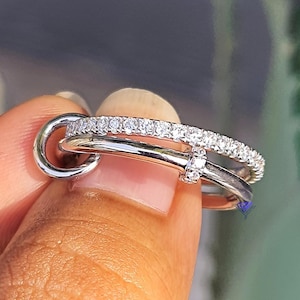 Könnte beinhalten: Ein silberfarbener Ring mit einem Doppelband-Design. Ein Band ist glatt und poliert, während das andere mit einer Reihe kleiner, klarer, funkelnder Steine besetzt ist. Die beiden Bänder sind an einer Seite durch ein kleines, rundes Glied verbunden.