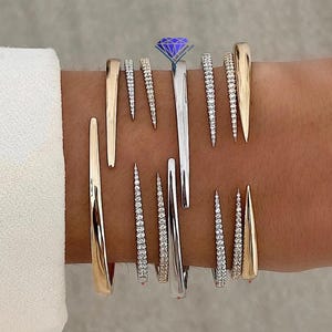 Puede incluir: Una colección de múltiples pulseras abiertas en la muñeca, con una mezcla de bandas de metal pulido en tono dorado y plateado. Algunas bandas son lisas y cónicas, mientras que otras están incrustadas con pequeñas piedras claras y brillantes.