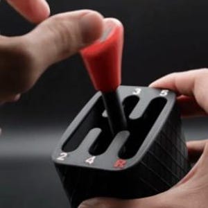 Pode incluir: Uma alavanca de mudanças preta com manípulo vermelho e placa de padrão de mudanças. A placa exibe os números brancos 2, 3, 4, 5 e a letra R vermelha, indicando as posições das mudanças para um veículo de transmissão manual.