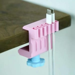 Peut inclure: Une pince organisatrice de câbles rose clair fixée sur une surface en bois. Le dispositif présente une série de fentes verticales pour maintenir les câbles et un bouton de serrage bleu clair en dessous pour fixer la pince sur le bord.
