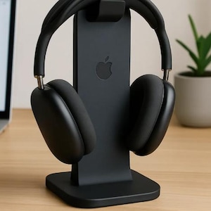 May include: A matte black headphone stand featuring a vertical support column with a debossed Apple logo, designed to hold over-ear headphones. The stand has a wide, stable rectangular base and a curved top hook to securely cradle the headband.