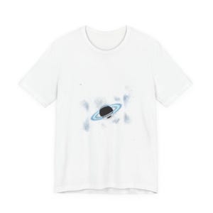 May include: White t-shirt featuring a black planet with a blue ring design. The graphic is centered on the front of the shirt, with a light blue and white splatter effect around the planet. The shirt has a crew neck.