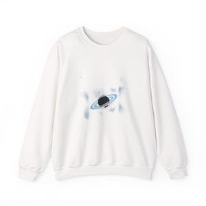 May include: A white crewneck sweatshirt featuring a black planet design with a light blue ring and cloud-like accents. The graphic is centered on the front of the long-sleeved top. The sweatshirt is made of a soft material.