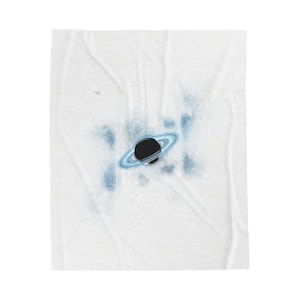 Pode incluir: Cobertor branco com um design de planeta negro. O planeta tem um anel azul claro e é rodeado por salpicos azuis, criando um padrão com tema espacial. O cobertor é feito de um material macio e texturizado.