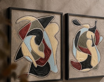 May include: Two rectangular framed wall art pieces featuring textured abstract shapes in a palette of black, deep red, olive green, and light blue against a neutral beige background. Each piece is enclosed in a thin black frame.