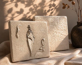 May include: Two textured, square, beige wall art pieces. One features an abstract relief design of a figure and organic shapes. The other is a plain square. The art pieces are on a beige fabric, with shadows from a plant in the background.