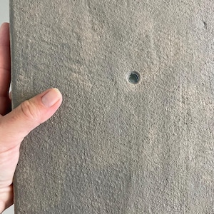 以下が含まれることがあります： 小さな穴のある、グレーのテクスチャ加工された長方形の物体。物体は手で持たれています。表面はざらざらとした不均一な外観で、コンクリートや石材を示唆しています。色はくすんだグレーです。