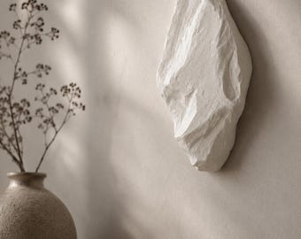May include: A white, irregularly shaped wall sculpture with a textured surface. The sculpture is mounted on a neutral-toned wall, creating a minimalist aesthetic. A vase with dried flowers and a black bowl are in the foreground.