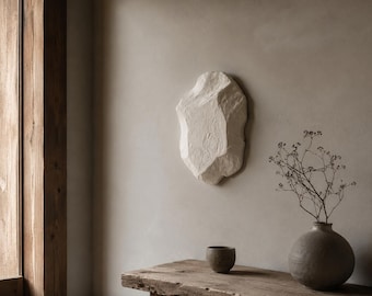 May include: A textured, off-white, rock-shaped wall sculpture. A small, dark brown ceramic bowl and a round, gray vase with dried branches sit on a weathered wooden table. The wall is a neutral beige color.