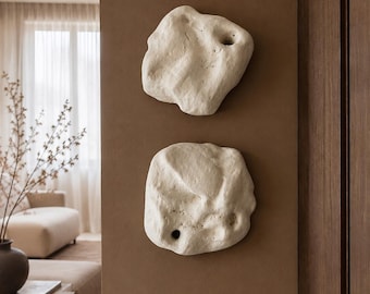 May include: Two off-white, irregularly shaped wall sculptures with porous, textured surfaces and small circular indentations. These sculptural pieces are mounted vertically on a flat, solid brown wall surface, displaying organic, stone-like forms.