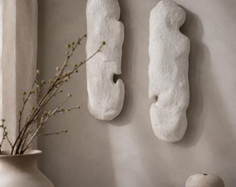May include: Two white, abstract wall sculptures with a textured surface, each with a curved cutout. The sculptures are mounted on a neutral-colored wall. A vase with branches, a small bowl, and books are on a wooden surface.