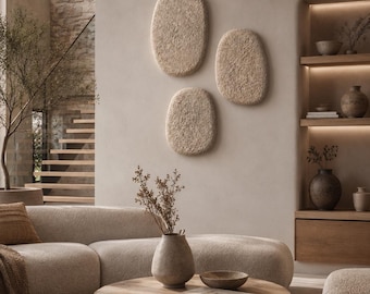 May include: Three oval-shaped, textured beige wall art pieces mounted on a neutral wall above a curved sofa. A round wooden coffee table holds a ceramic vase with dried stems, a small wooden bowl, and a book, with a large dome pendant light above.