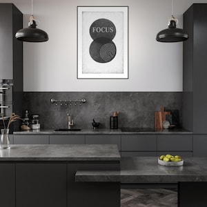 Könnte beinhalten: Eine moderne Küche mit dunkelgrauen Schränken und Arbeitsplatten. Ein gerahmter Druck an der Wand zeigt das Wort "FOCUS" über einer Grafik aus zwei sich überlappenden Kreisen. Schwarze Pendelleuchten hängen über der Kücheninsel.