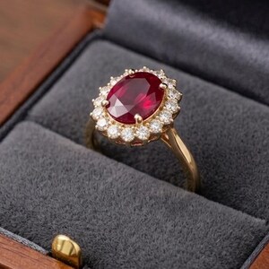 May include: A gold ring featuring a large, oval-cut deep red ruby at the centre, surrounded by a halo of small, round, clear white diamonds. The ring is displayed inside a dark wooden box with a soft, dark grey velvet interior lining.