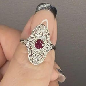 Può includere: Anello color argento con un design a filigrana ornato. L'anello presenta una gemma rossa rotonda centrale, circondata da pietre chiare più piccole. Il design dell'anello è simmetrico, in stile vintage o Art Déco.