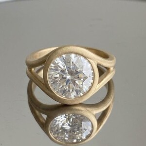 Könnte beinhalten: Ein goldener Ring mit einem großen, runden, klaren Edelstein. Der Ring hat ein einzigartiges Design mit einem wirbelnden Band, das den Edelstein umschließt. Der Ring spiegelt sich in der glänzenden Oberfläche darunter.
