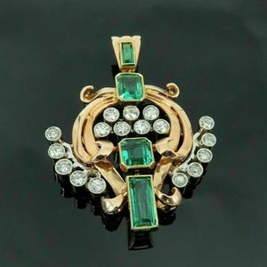 Könnte beinhalten: Goldfarbener Anhänger mit smaragdgrünen Edelsteinen im Smaragdschliff und kleinen, runden, klaren Edelsteinen. Der Anhänger hat ein verziertes Design mit geschwungenen Elementen und einen dunklen Hintergrund.