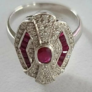 Könnte beinhalten: Silberring mit einem zentralen ovalen Rubin und Diamanten. Der Ring hat ein geometrisches Design mit Rubinakzenten. Das Band ist aus poliertem Silber.