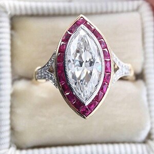 Op de afbeelding: Een marquise-geslepen diamanten ring met een halo van kleine, vierkant geslepen robijnen. De ring heeft een gouden band met ingewikkelde details en kleine accentdiamanten op de schouders. De ring zit in een witte doos.