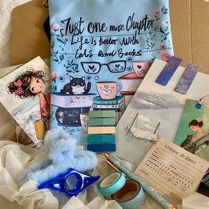 Peut inclure: Un ensemble d'accessoires de lecture et de papeterie, dont une housse de livre bleu clair avec le texte "Just one more Chapter, Life is better with Cats and Books". L'image comprend également des marque-pages, des stylos et une lampe de lecture.
