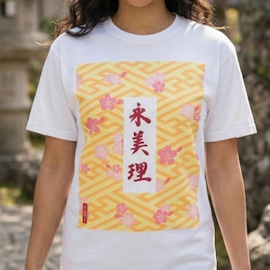 May include: White t-shirt featuring a rectangular design with a yellow geometric pattern and pink cherry blossoms. The design includes Japanese characters in red. A small red text is at the bottom of the design.