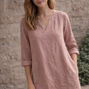 Puede incluir: Vestido de lino color rosa empolvado con cuello en V y mangas tres cuartos. El vestido tiene un corte holgado, bolsillos y un dise&ntilde;o sencillo y elegante. La modelo lleva un delicado collar de oro.