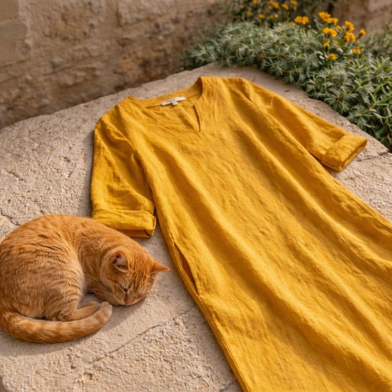 Puede incluir: Un vestido de lino amarillo mostaza con cuello en V y bolsillos laterales. El vestido tiene mangas tres cuartos y est&aacute; sobre una superficie de piedra. Un gato atigrado naranja est&aacute; acurrucado junto al vestido.