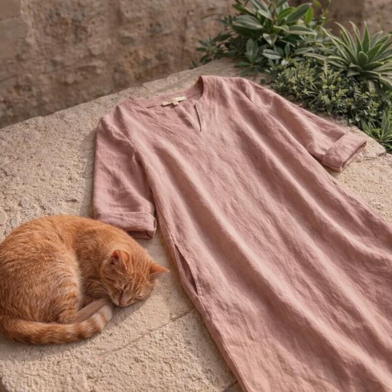 Puede incluir: Un vestido de lino rosa empolvado con cuello en V y mangas hasta el codo descansa sobre una superficie de piedra junto a un gato atigrado naranja durmiendo. El vestido tiene un corte holgado y una abertura lateral. Hay plantas verdes en el fondo.