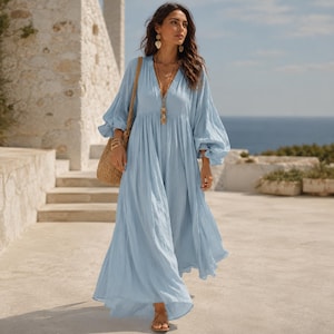 K&ouml;nnte beinhalten: Ein hellblaues Maxikleid mit langen, volumin&ouml;sen &Auml;rmeln, einem tiefen V-Ausschnitt und einer gerafften Taille. Das Kleid hat einen flie&szlig;enden, bodenlangen Rock und wird mit einer gewebten Stroh-Umh&auml;ngetasche und goldenen Ohrh&auml;ngern kombiniert.
