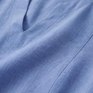 Puede incluir: Primer plano de una blusa de lino azul claro con cuello en V. La tela tiene una apariencia texturizada con costuras visibles a lo largo del escote y las costuras. La blusa est&aacute; hecha de un material ligero.
