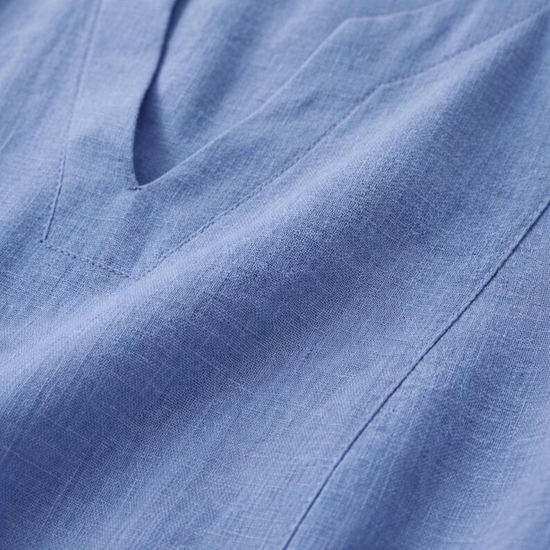 Puede incluir: Primer plano de una blusa de lino azul claro con cuello en V. La tela tiene una apariencia texturizada con costuras visibles a lo largo del escote y las costuras. La blusa est&aacute; hecha de un material ligero.