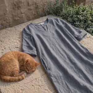 Puede incluir: Un vestido de lino azul claro con cuello en V y bolsillos laterales. El vestido est&aacute; sobre una superficie de piedra junto a un gato atigrado naranja durmiendo. El vestido tiene mangas cortas.