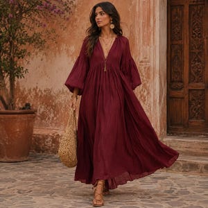 K&ouml;nnte beinhalten: Ein tiefburgunderrotes Maxikleid mit tiefem V-Ausschnitt, langen, volumin&ouml;sen Bischofs&auml;rmeln mit gerafften B&uuml;ndchen und einem flie&szlig;enden, bodenlangen Rock. Das Kleid wird mit einer gewebten beigen Tragetasche und braunen Riemchensandalen kombiniert.