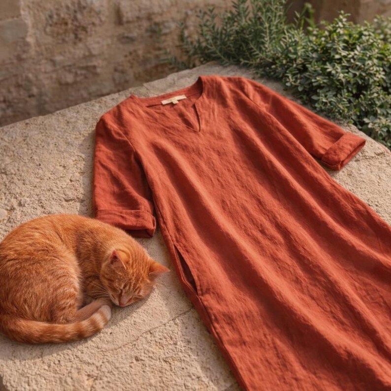 Puede incluir: Vestido de lino color &oacute;xido con cuello en V y bolsillos laterales. El vestido tiene mangas hasta el codo. Un gato atigrado naranja est&aacute; acurrucado dormido junto al vestido sobre una superficie de piedra.