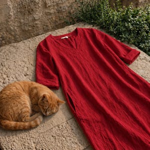 Puede incluir: Un vestido de lino rojo vibrante con cuello en V y mangas tres cuartos descansa sobre una superficie de piedra. El vestido tiene un corte holgado y un bolsillo lateral. Un gato atigrado naranja est&aacute; acurrucado durmiendo junto al vestido.
