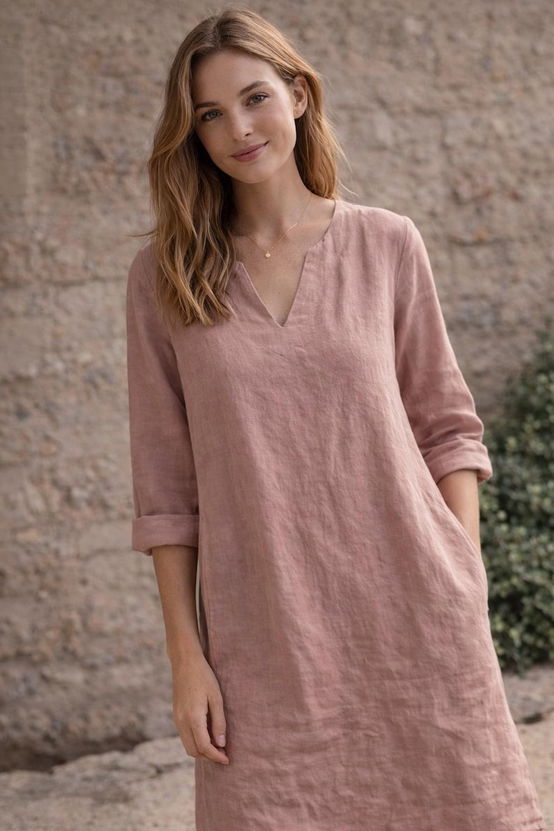 Puede incluir: Vestido de lino color rosa empolvado con cuello en V y mangas tres cuartos. El vestido tiene un corte holgado, bolsillos y un dise&ntilde;o sencillo y elegante. La modelo lleva un delicado collar de oro.