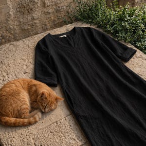 Puede incluir: Un vestido negro de lino con cuello en V y mangas tres cuartos descansa sobre una superficie de piedra. Un gato atigrado naranja est&aacute; acurrucado durmiendo junto al vestido. El vestido tiene un corte holgado y una abertura lateral.