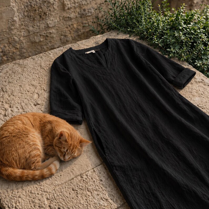 Puede incluir: Un vestido negro de lino con cuello en V y mangas tres cuartos descansa sobre una superficie de piedra. Un gato atigrado naranja est&aacute; acurrucado durmiendo junto al vestido. El vestido tiene un corte holgado y una abertura lateral.