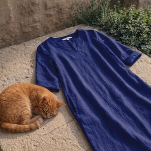 Puede incluir: Un vestido largo azul marino con cuello en V y mangas tres cuartos descansa sobre una superficie de piedra. Un gato atigrado naranja est&aacute; acurrucado durmiendo junto al vestido. El vestido parece estar hecho de una tela ligera.